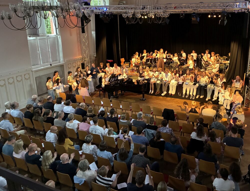 Voll besetzte Aula des HErder-Gymnasiums und alle Musiker*innen auf der Bühne beim Sommerkonzert 2026