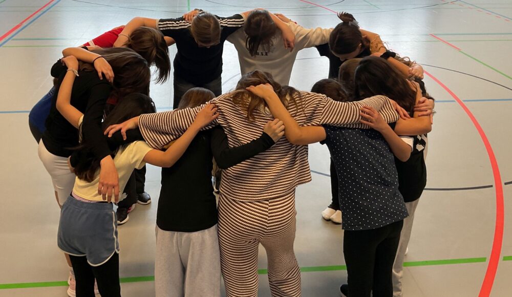 De Mitgliederinnen der Tanz-AG Stehen in einem Kreis und haben die Arme gegenseitig über die Schultern gelegt.